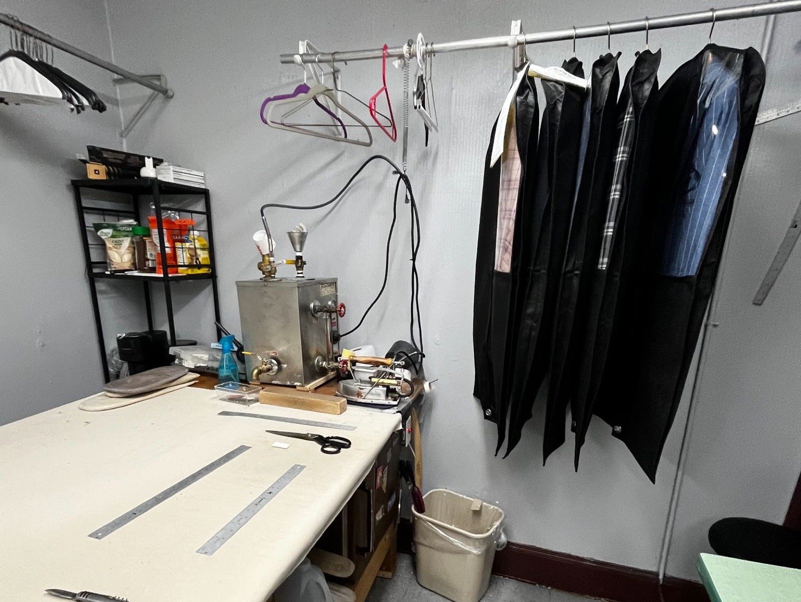Cutting table with rulers and shears beside a steam pressing iron and finished shirts in garment bags